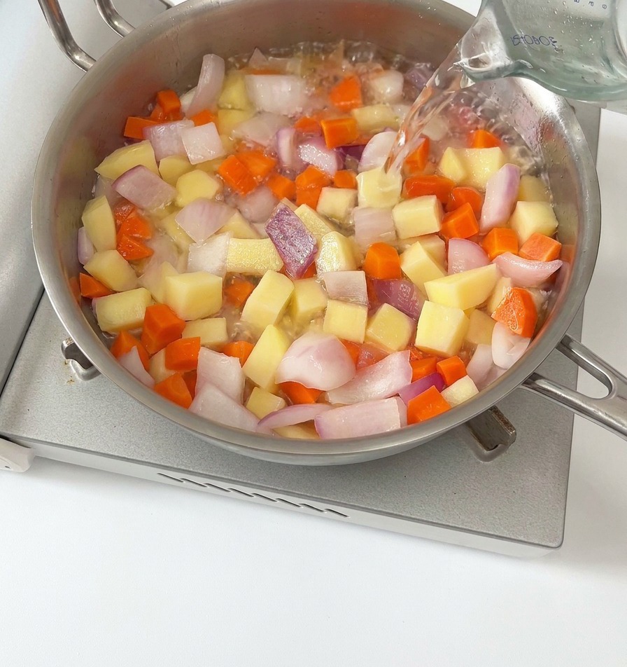 Water being poured from a glass measuring cup into a pan filled with diced carrots, potatoes, and red onions.