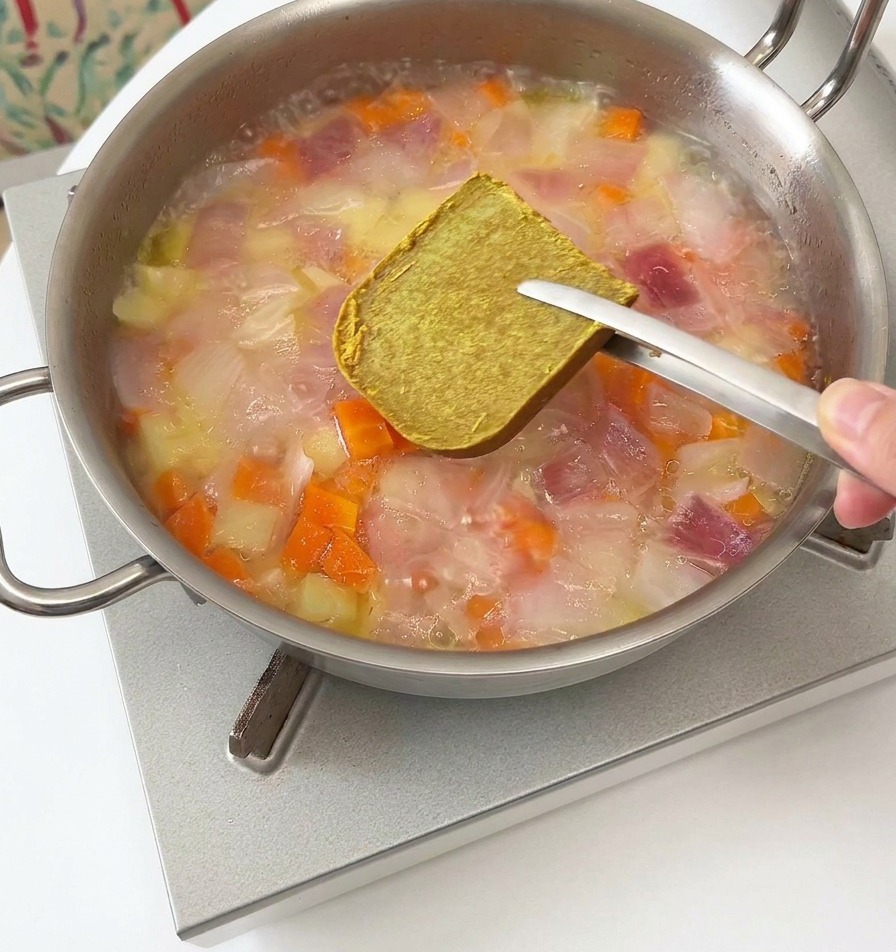 Metal tongs holding a square, solid brown curry roux cube over a steaming pan of vegetables and water.