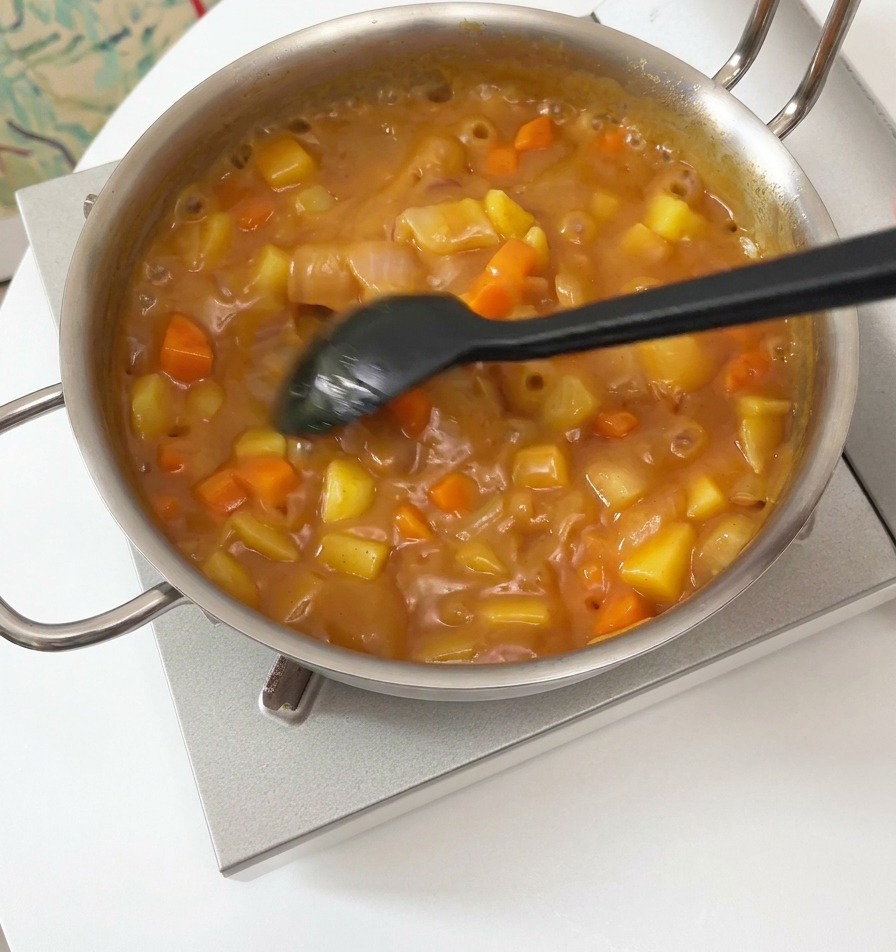 A black spatula stirring a thick, bubbling brown curry sauce with visible chunks of potato and carrot in a silver pot.