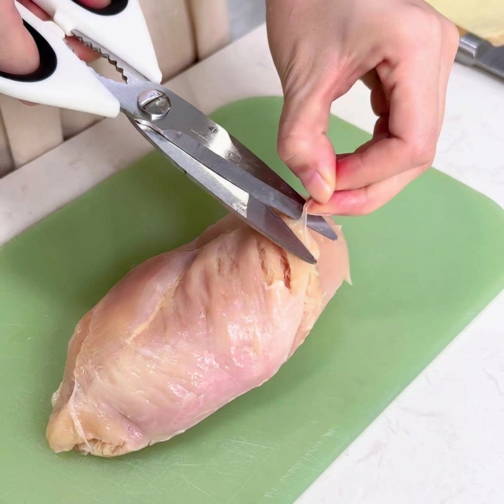 A hand using white kitchen shears to pull and snip away the thin, translucent connective tissue from a raw chicken breast on a green cutting board.