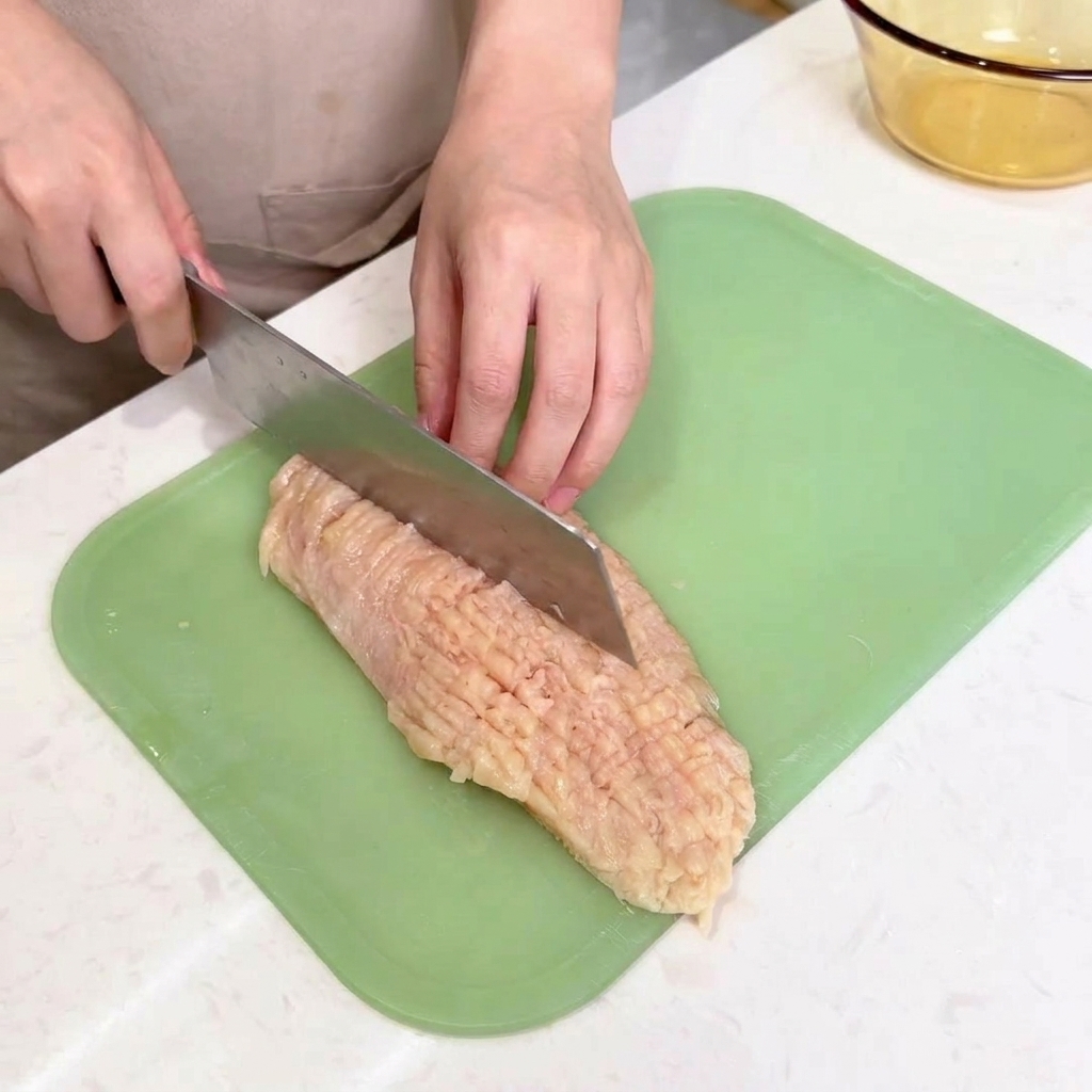 The blunt spine of a large chefs knife being used to strike and tenderize a flat piece of raw chicken on a cutting board.