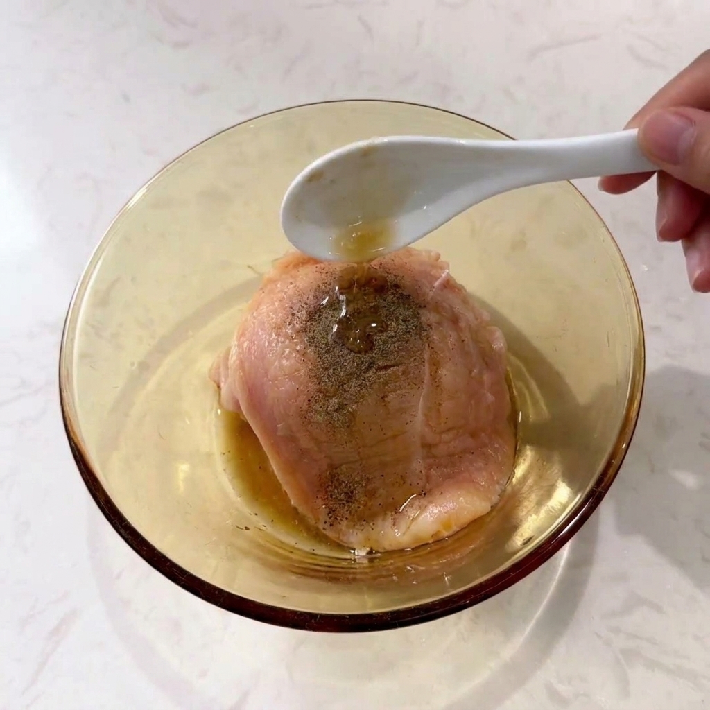 A spoon pouring liquid seasoning over a raw chicken breast topped with ground black pepper in a clear glass bowl.