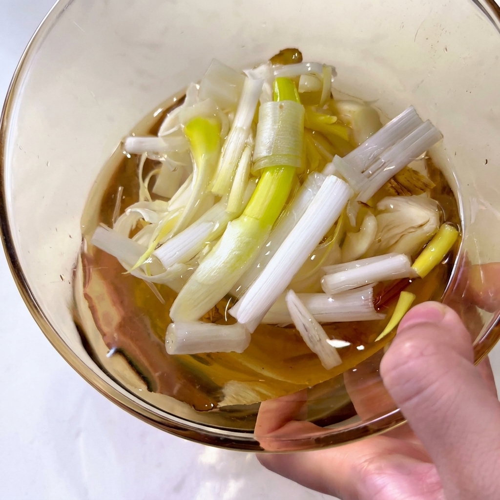 A close-up of a glass bowl filled with clear liquid and large pieces of chopped green scallions and sliced ginger.