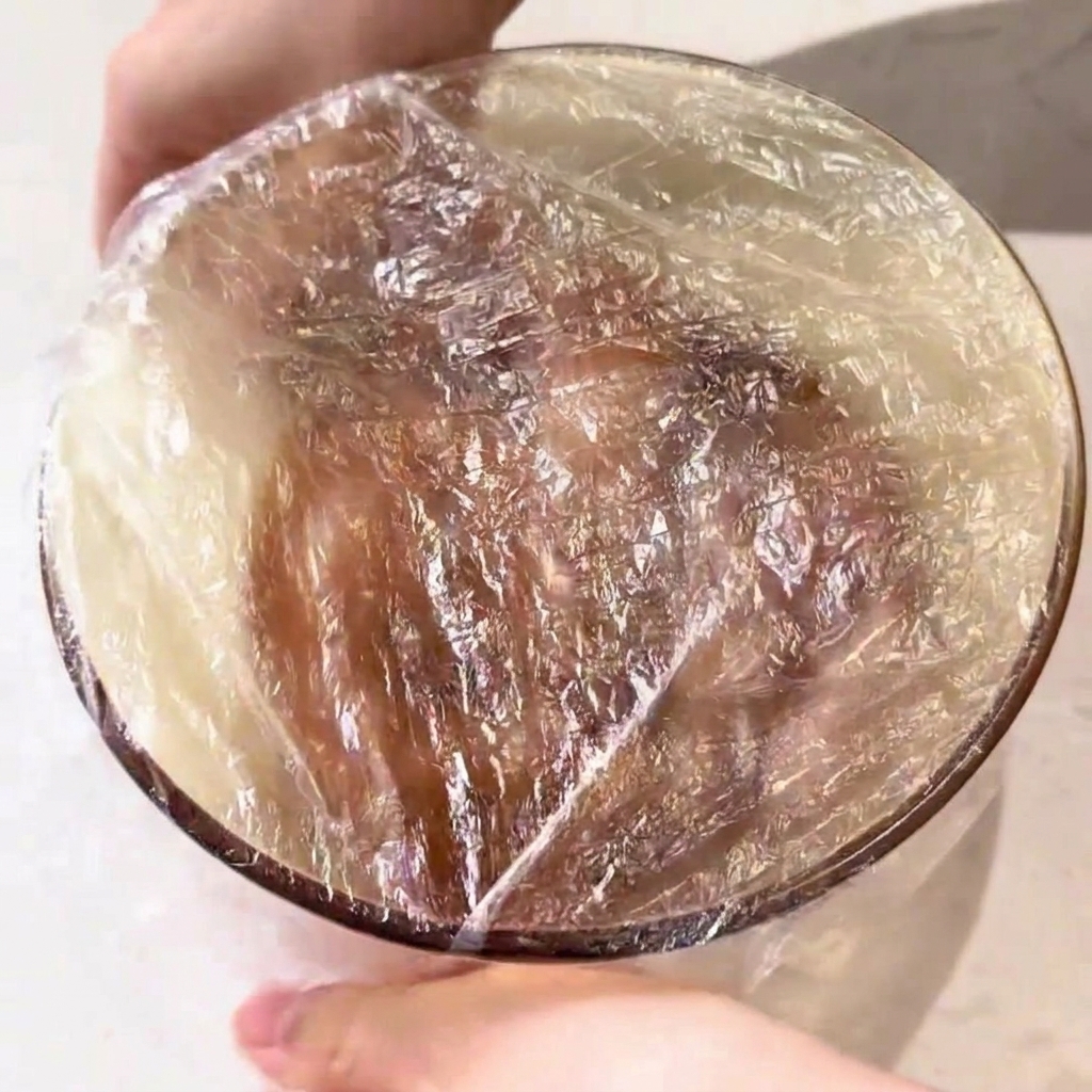 A glass bowl containing marinated chicken pieces, tightly sealed with a layer of transparent plastic wrap.