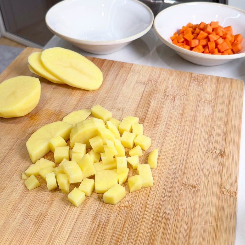 Diced yellow potato cubes piled on a wooden cutting board next to a bowl of already diced carrots.