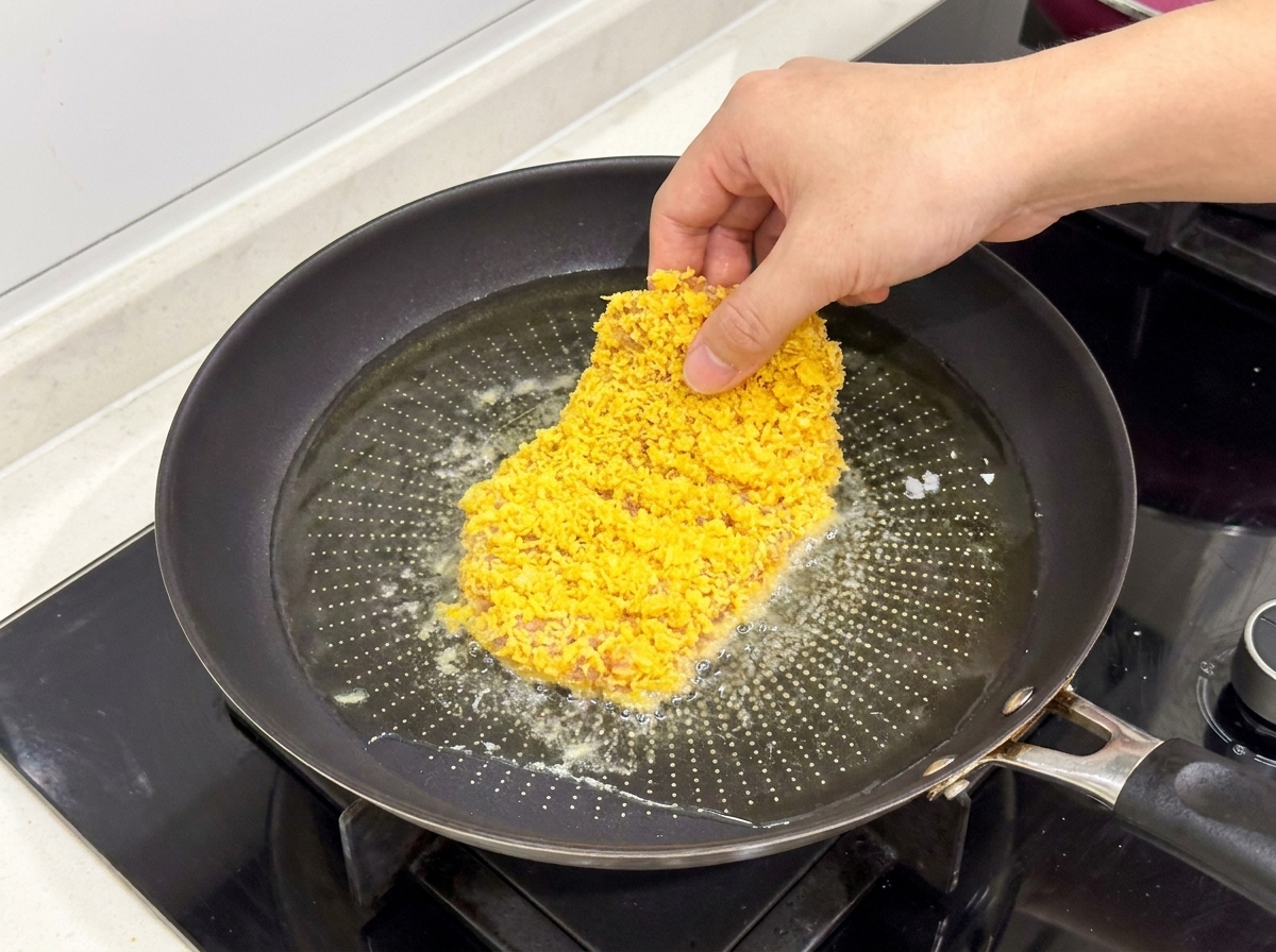 A breaded chicken cutlet being placed into a frying pan with shimmering hot oil.