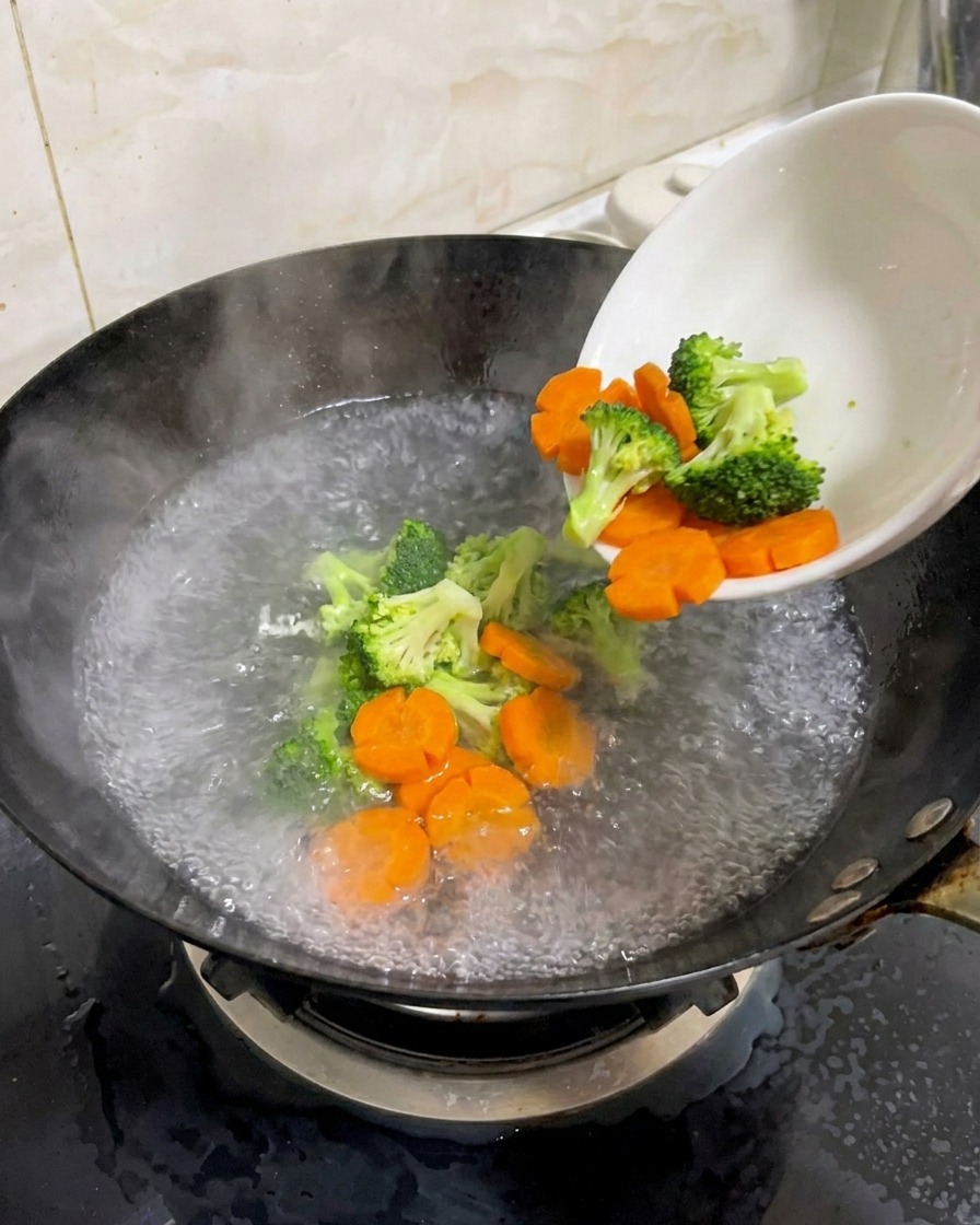 Fresh broccoli and flower-shaped carrots being poured from a white bowl into a wok of boiling water.