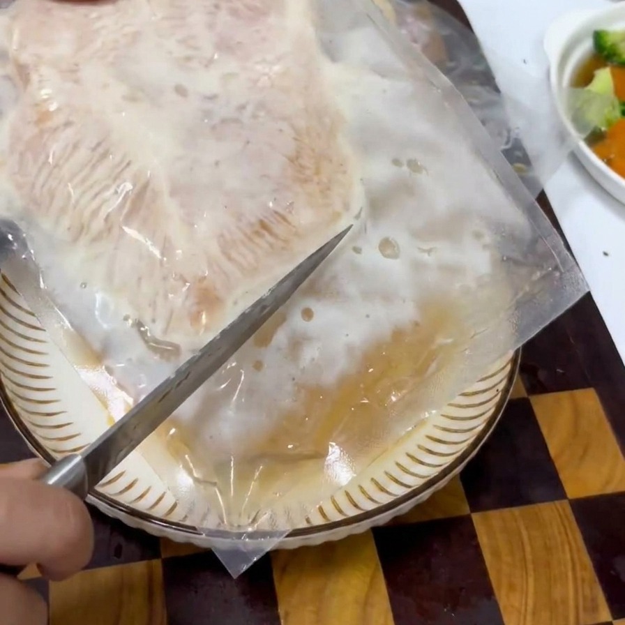 A hand using a knife to slice open a clear plastic vacuum pack containing a piece of marinated chicken meat.