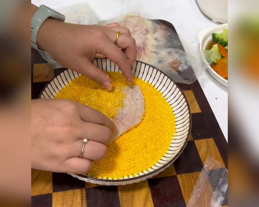 Hands pressing a marinated chicken breast into a shallow white bowl filled with bright yellow breadcrumbs.