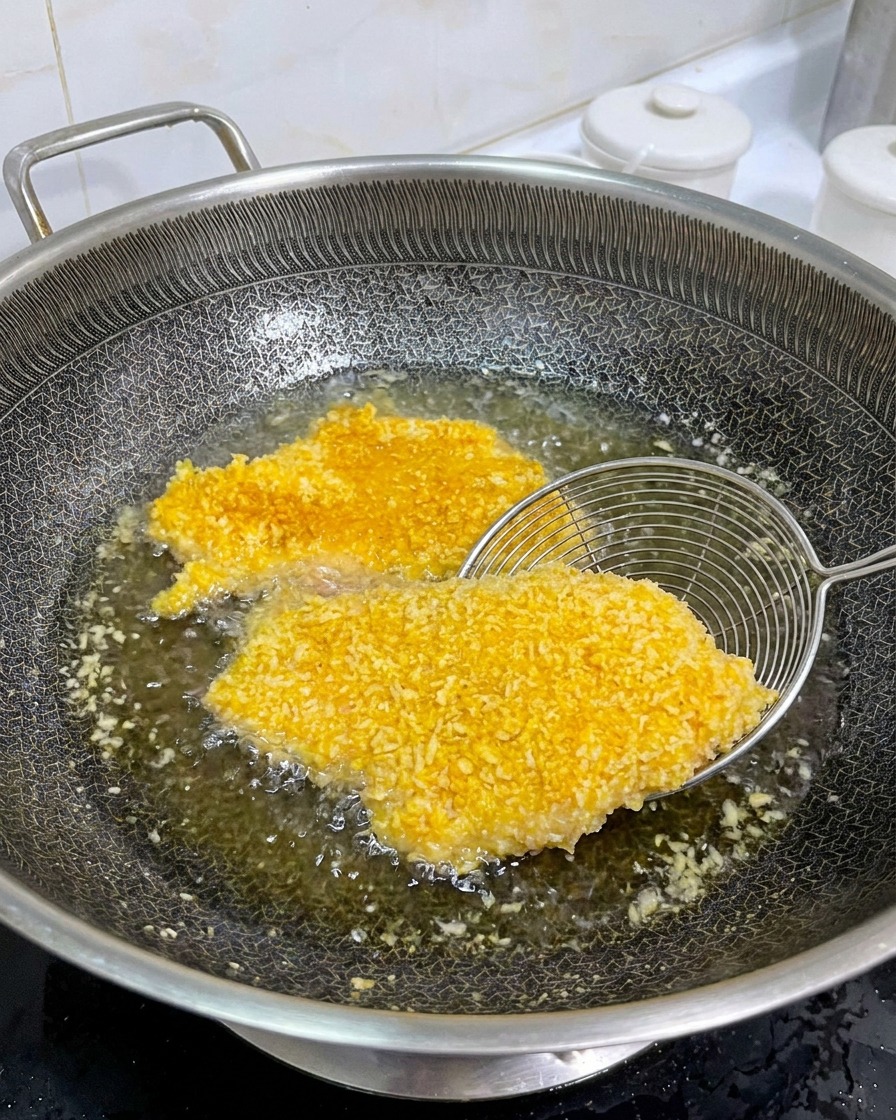 A metal strainer lifting a golden-brown, breaded chicken cutlet in a pan of hot oil to show the cooked side.