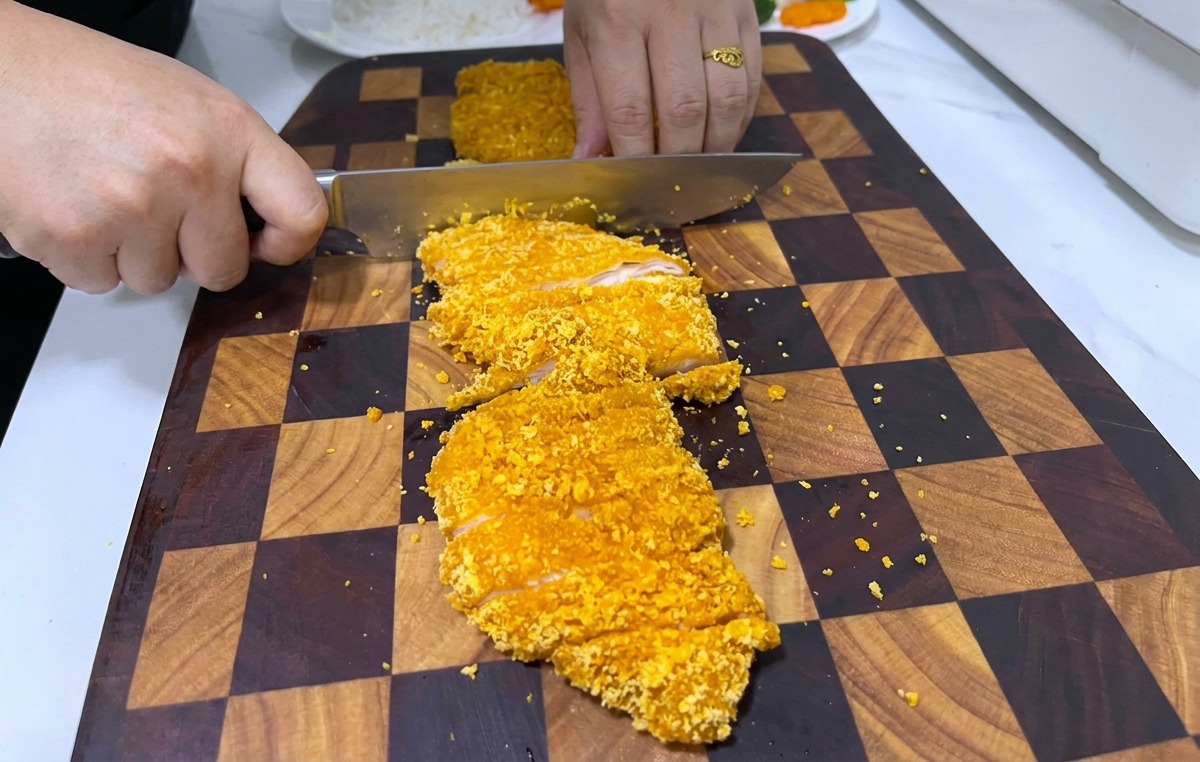 A person using a large knife to slice a crispy, breaded golden chicken steak on a dark wooden checkerboard cutting board.