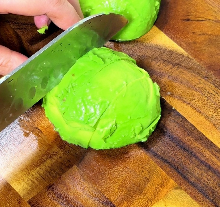 A close-up view of hands using a professional knife to dice a bright green peeled avocado into even cubes on a wooden surface.