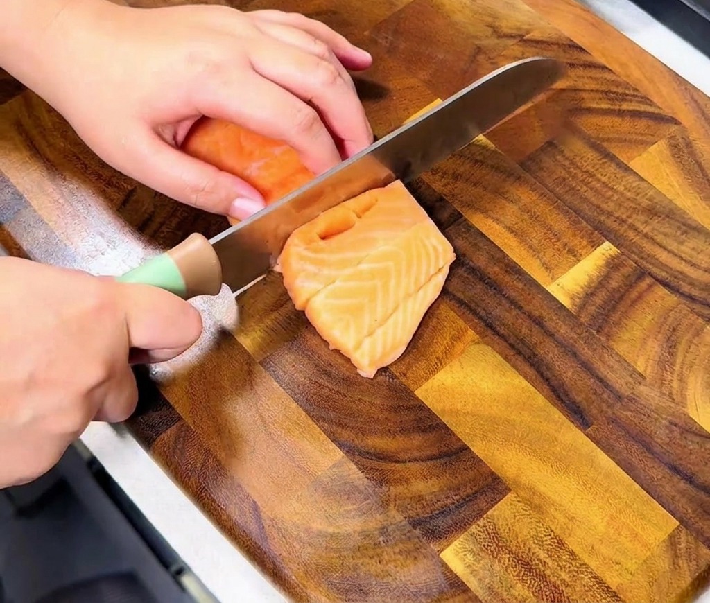 Hands slicing through a fresh orange salmon fillet on a wooden board to create uniform, bite-sized cubes.