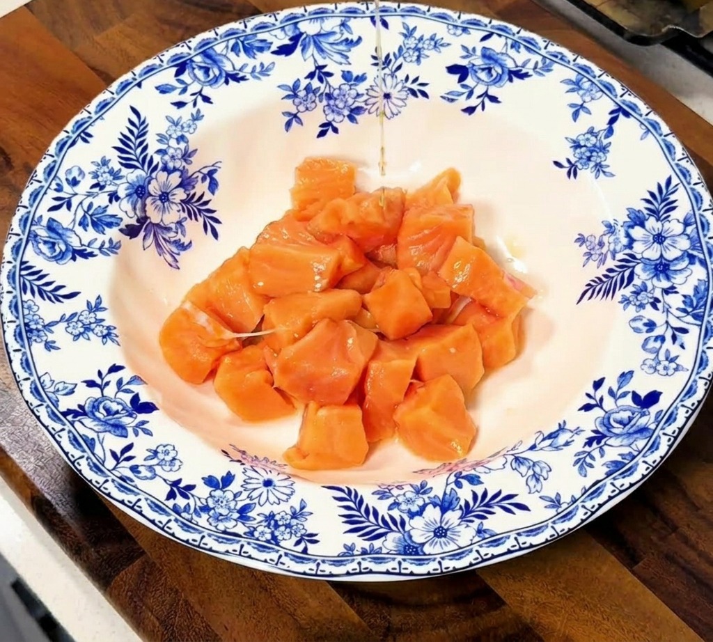 A stream of clear and dark liquid marinade being poured over a pile of raw salmon cubes in a floral-patterned ceramic bowl.