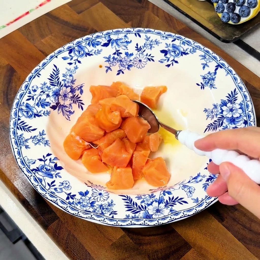 A hand using a white spoon to stir salmon cubes in a blue and white bowl, ensuring they are well-coated in the marinade liquid.