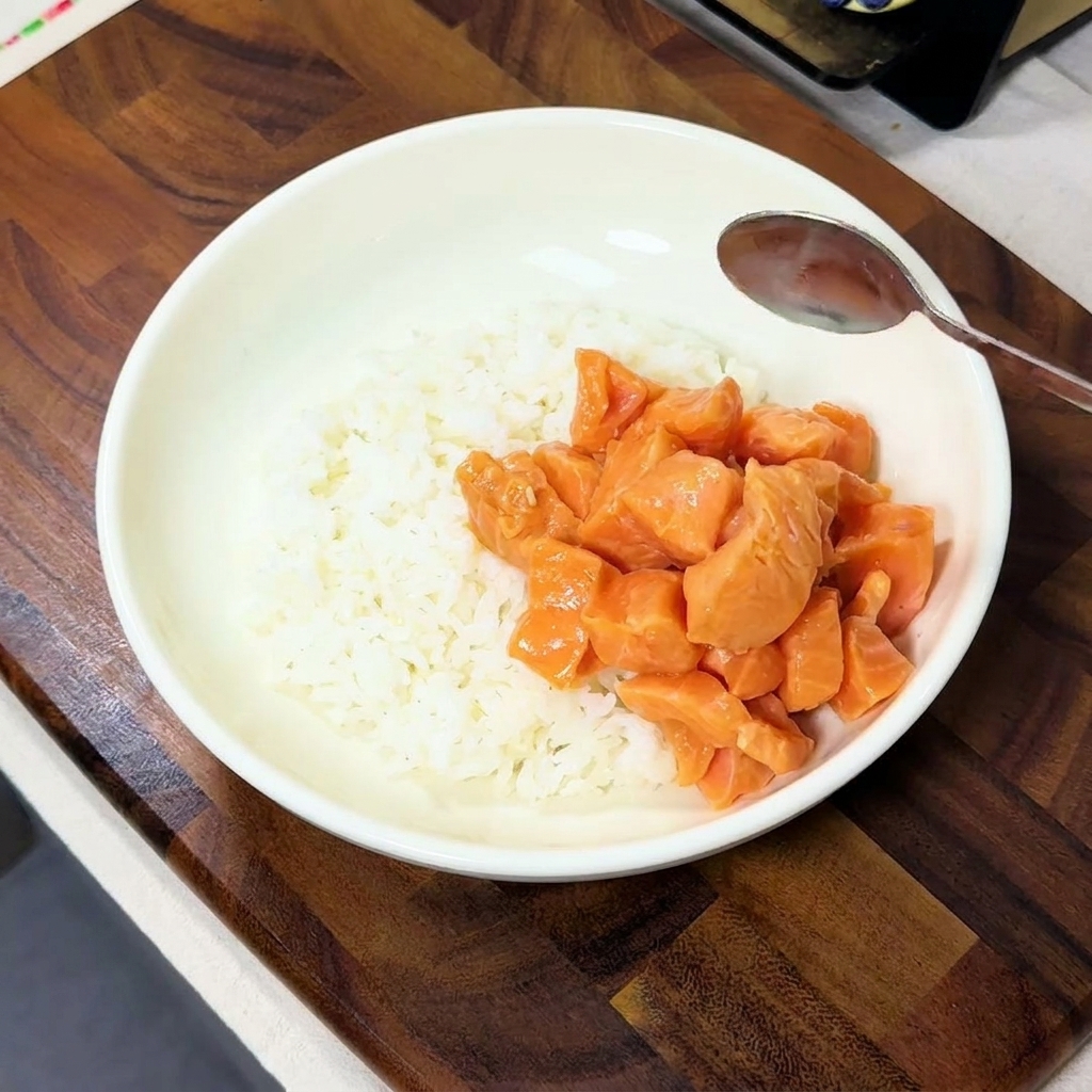 Marinated salmon cubes being spooned onto one side of a white bowl filled with steamed white rice.