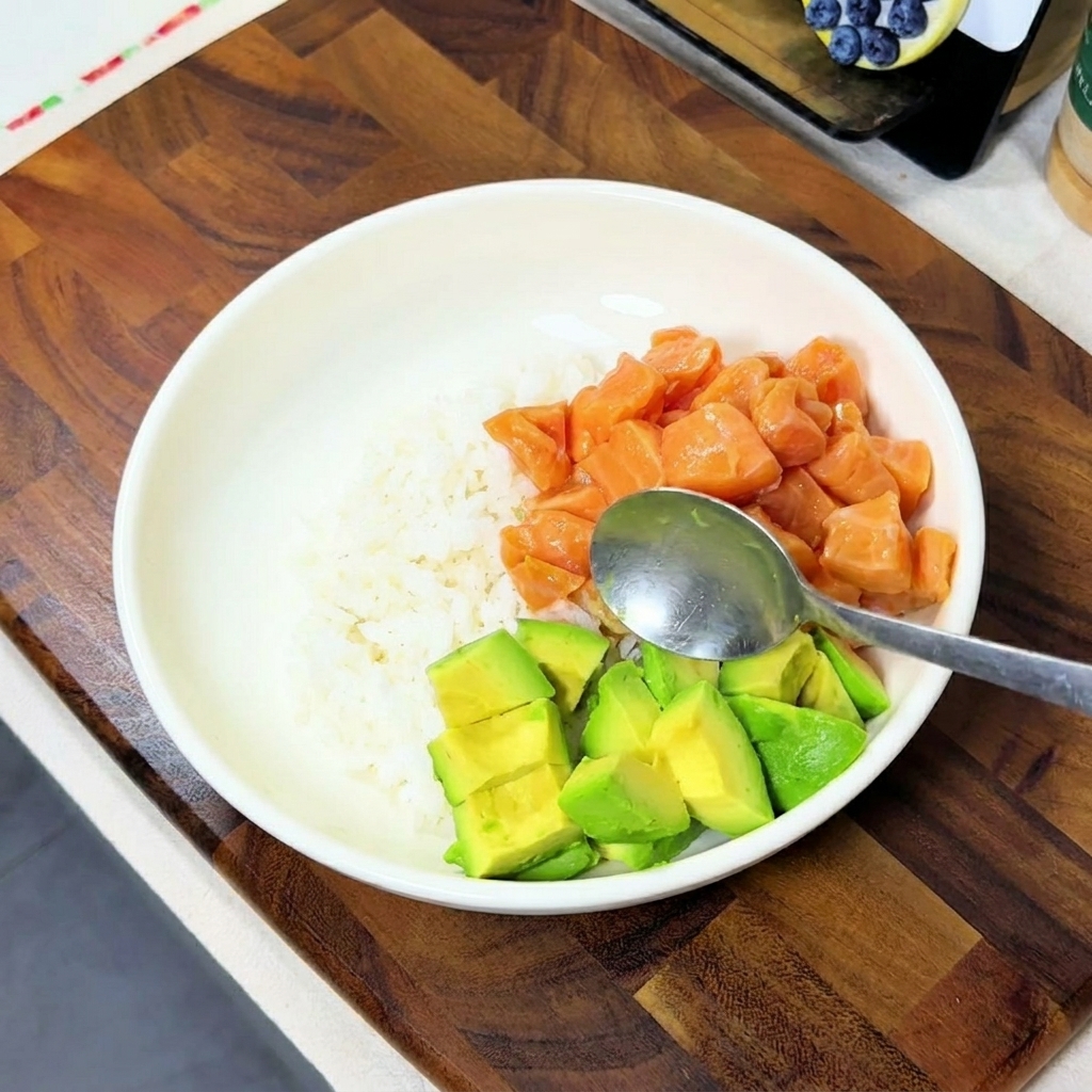 Avocado cubes being added with a spoon into a white bowl that already contains rice and marinated salmon.