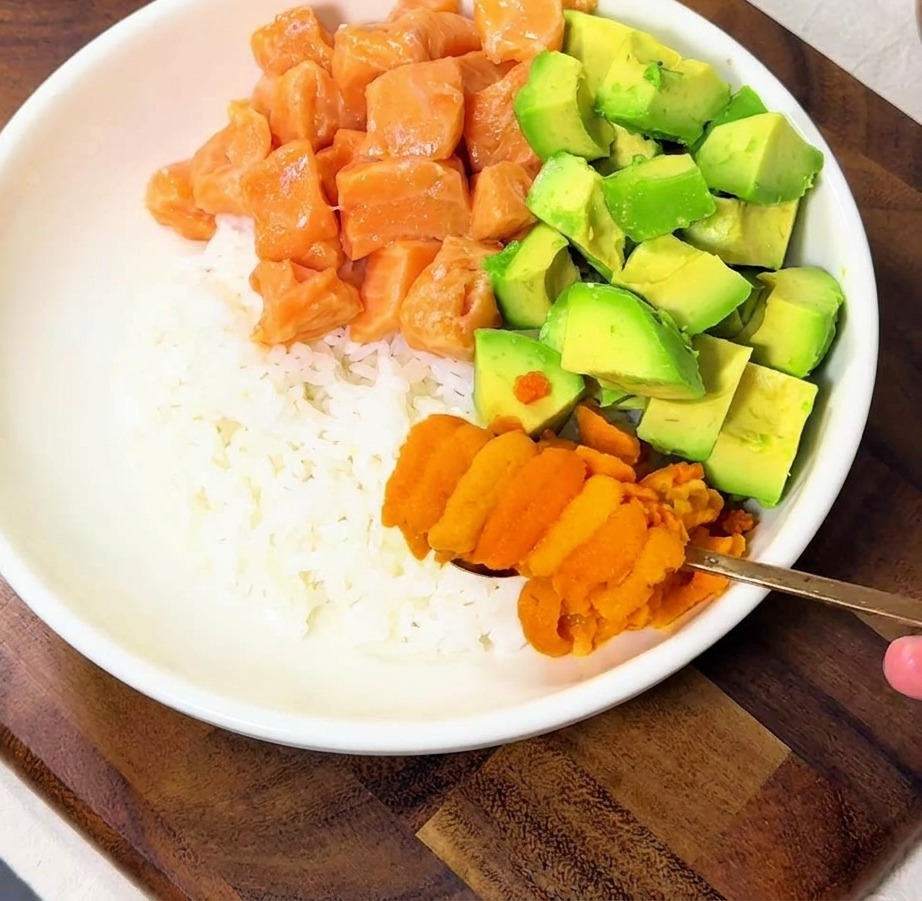 A hand using a spoon to place bright orange sea urchin pieces onto a bowl of rice topped with salmon and avocado cubes.