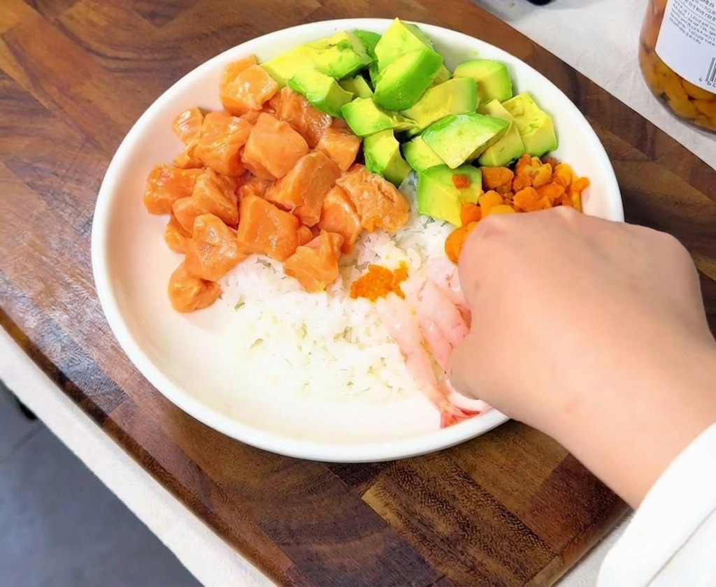 A hand arranging several translucent pink sweet shrimp on the side of the rice bowl.