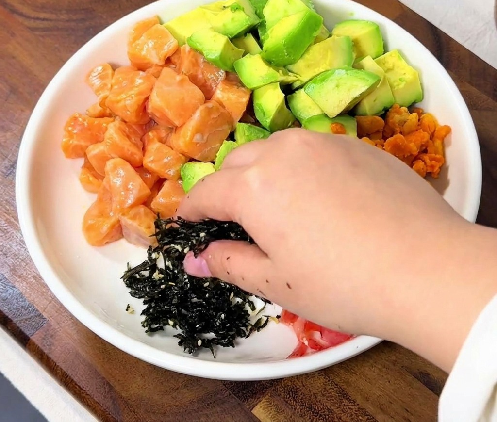 A hand sprinkling a pile of black shredded seaweed onto the rice bowl next to the salmon cubes.