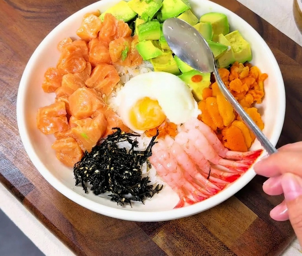 A spoon lowering a white onsen egg into the middle of the colorful seafood toppings in the bowl.