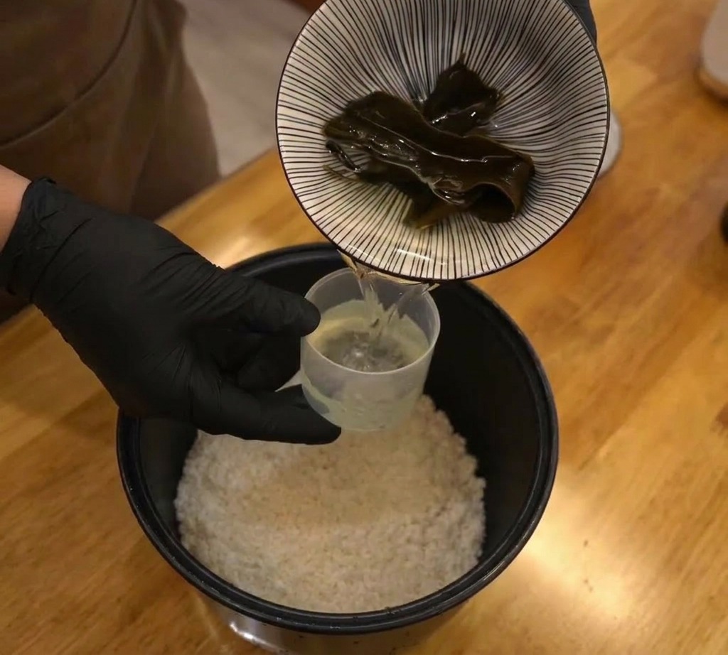 A dark piece of dried kombu being placed into a rice cooker pot filled with uncooked white rice and water.