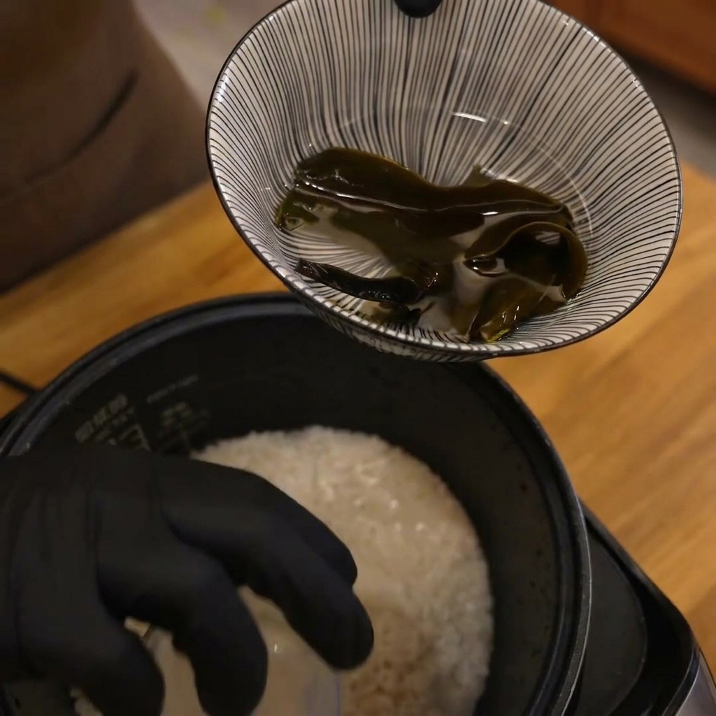 A gloved hand holding a small measuring cup over a pot of uncooked white rice, preparing to add water.