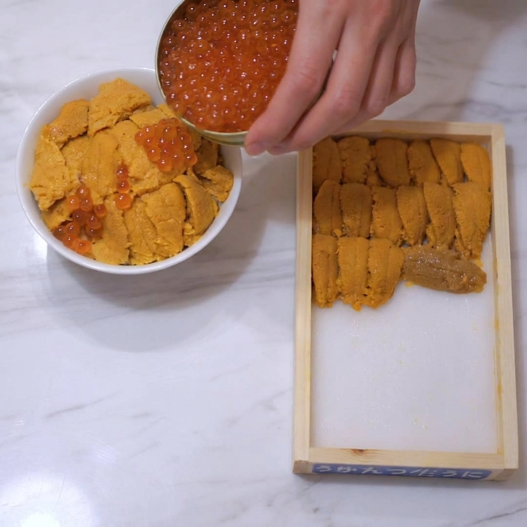 Shiny orange-red salmon roe being poured from a small gold tin onto a bowl topped with sea urchin.