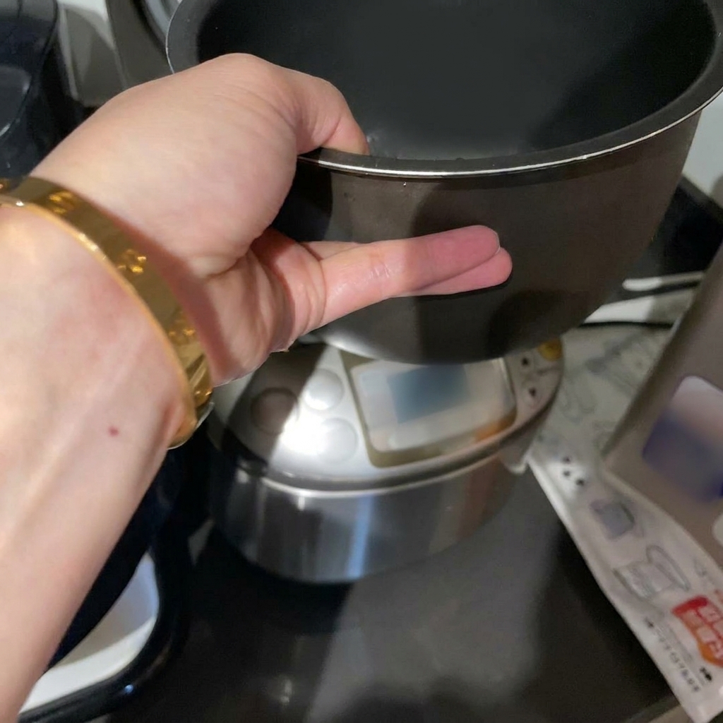 A hand holding a dark rice cooker inner pot containing raw white rice submerged in water.