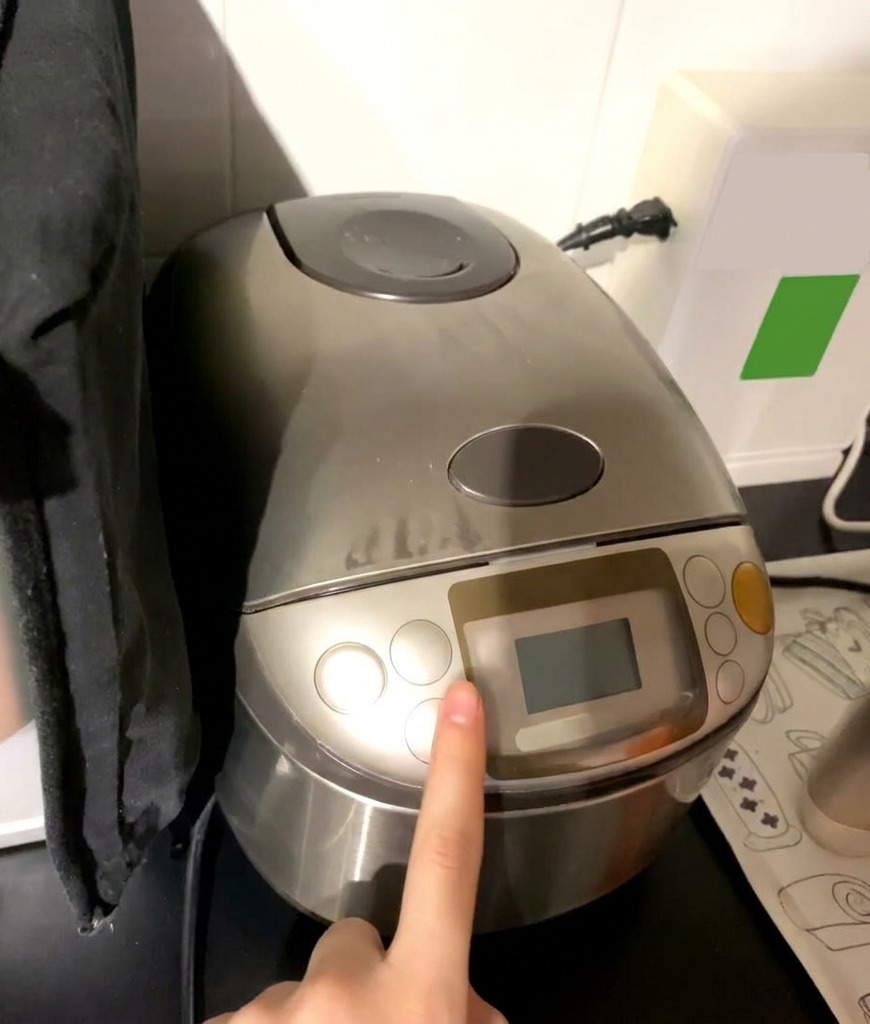 A finger pressing the start button on a silver Zojirushi rice cooker with the digital display visible.