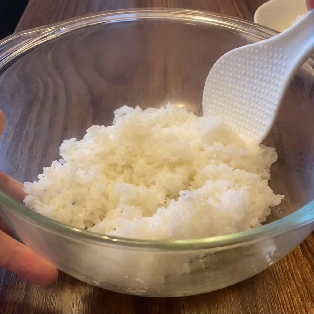 A white textured rice paddle fluffing steaming hot white rice inside a large clear glass mixing bowl.