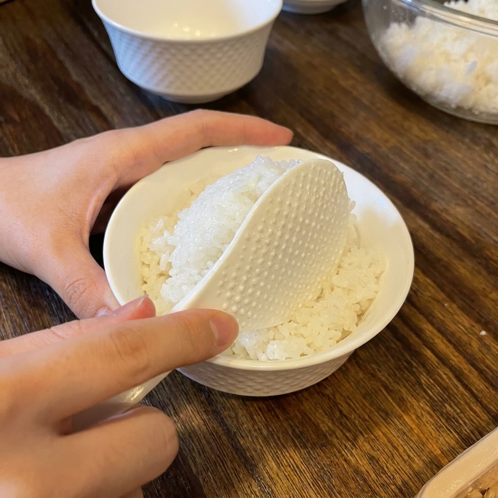 A white rice paddle placing a rounded scoop of seasoned white rice into a small textured white ceramic bowl.
