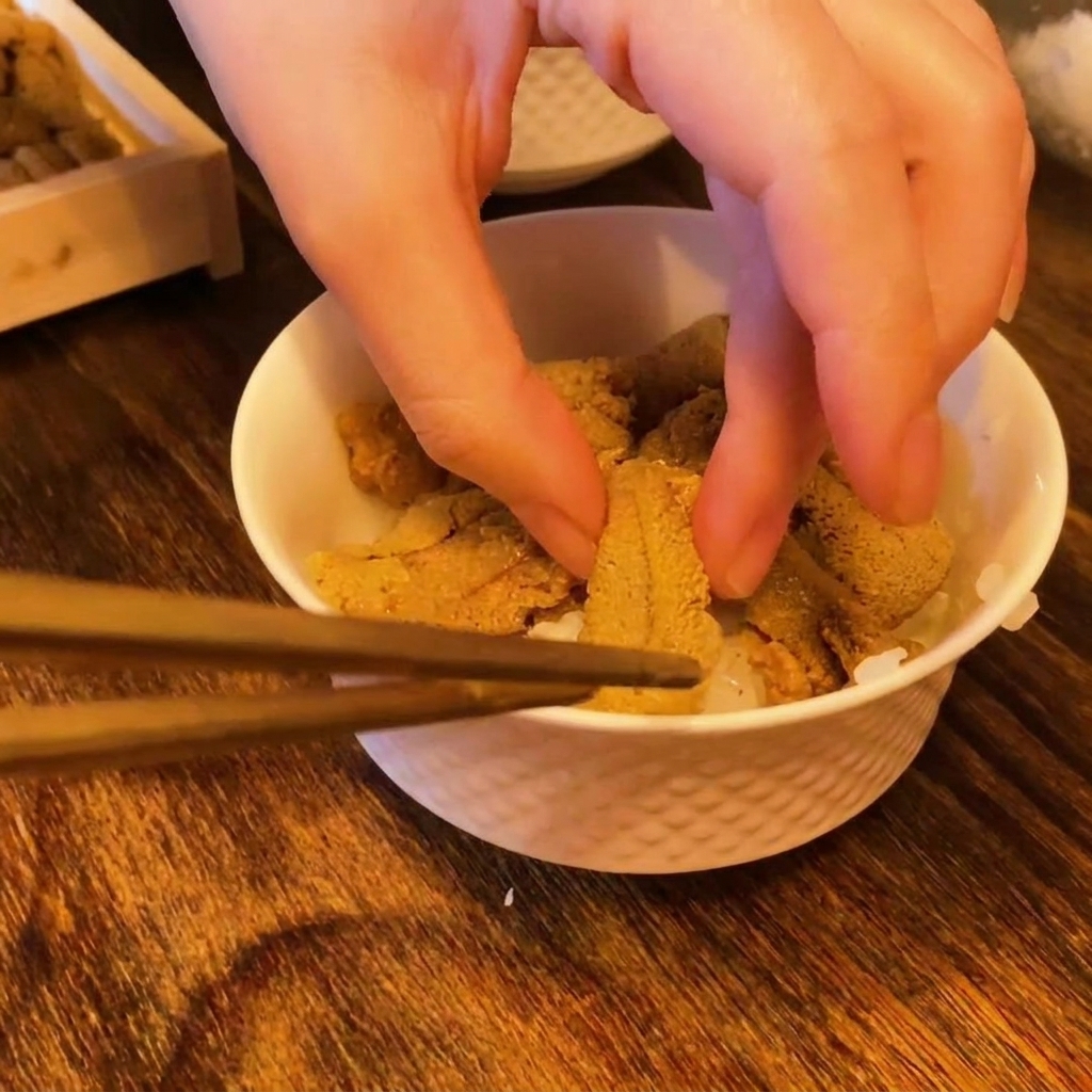 Chopsticks and fingers placing bright orange pieces of fresh sea urchin onto a bed of white sushi rice in a bowl.