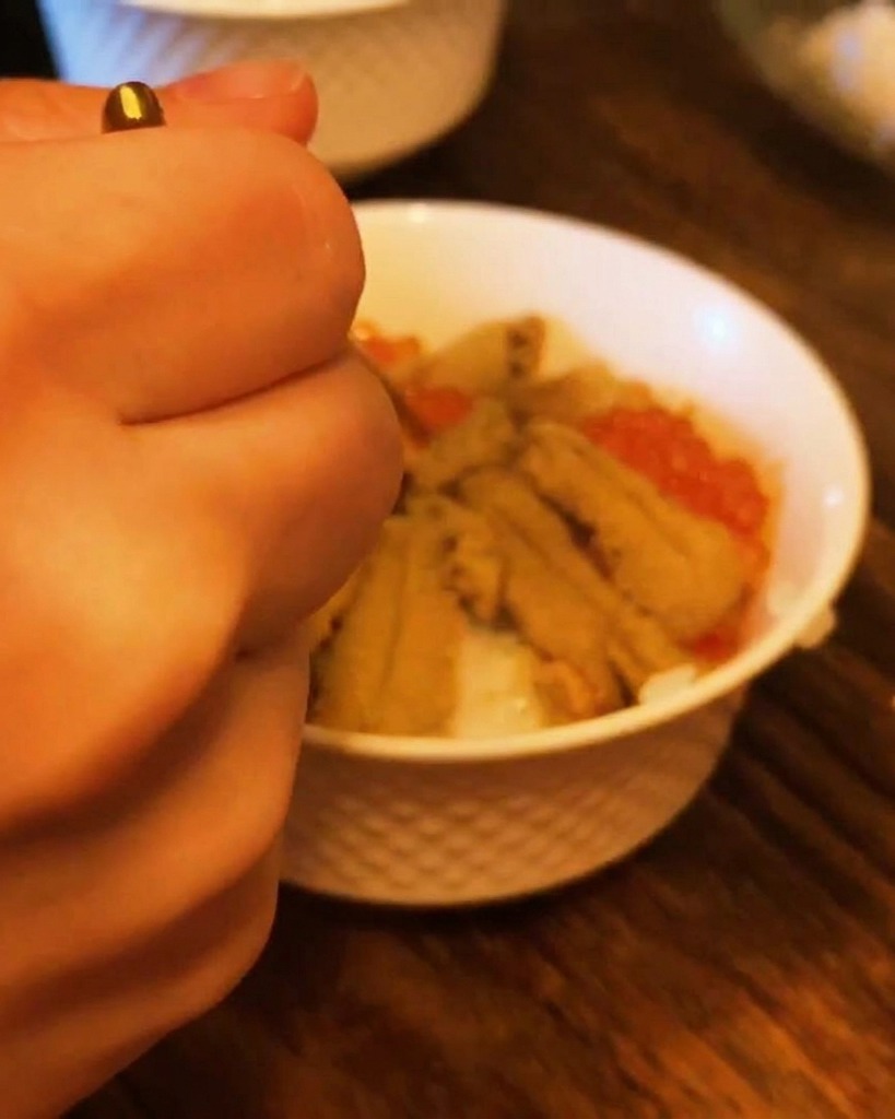 A close-up of a hand using a small spoon to fill the spaces in a sea urchin bowl with bright orange salmon roe.