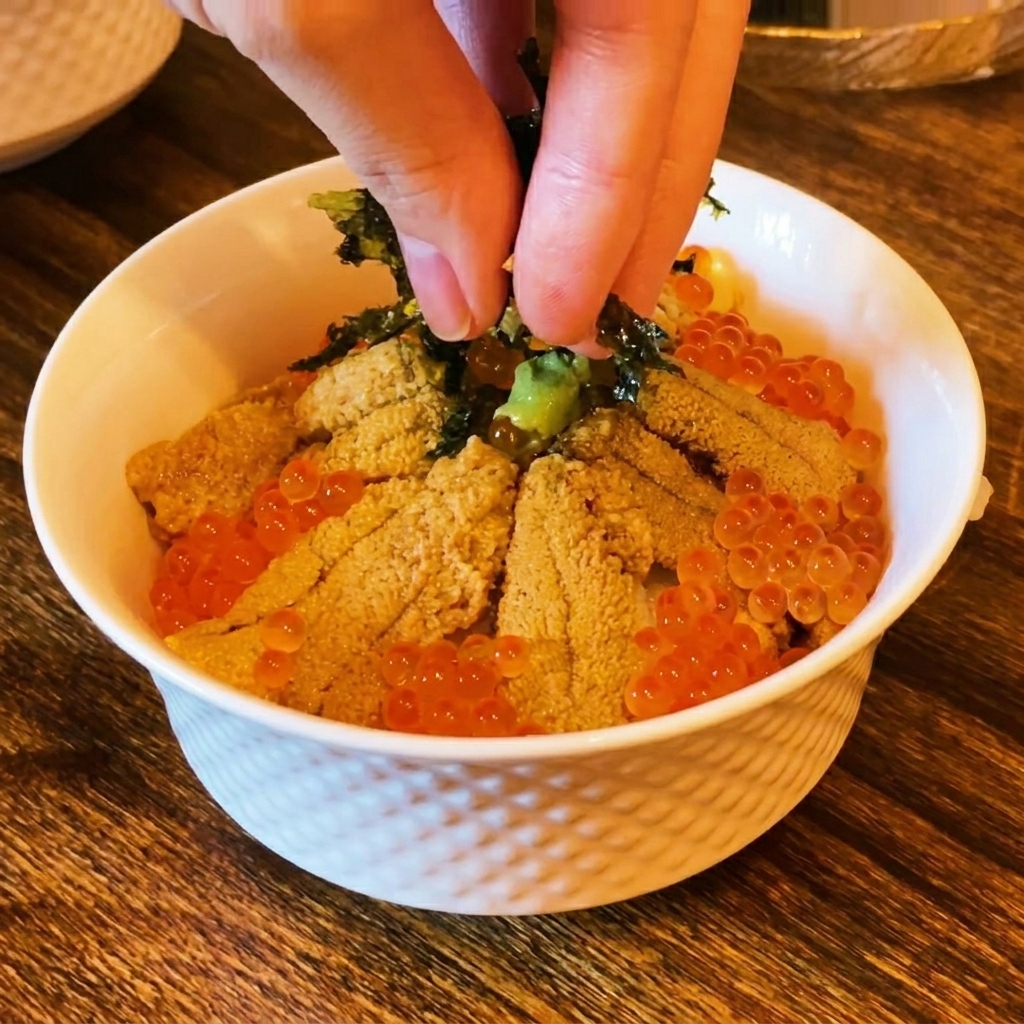 Fingers sprinkling dark shredded seaweed over the top of a sushi bowl filled with sea urchin and orange salmon roe.