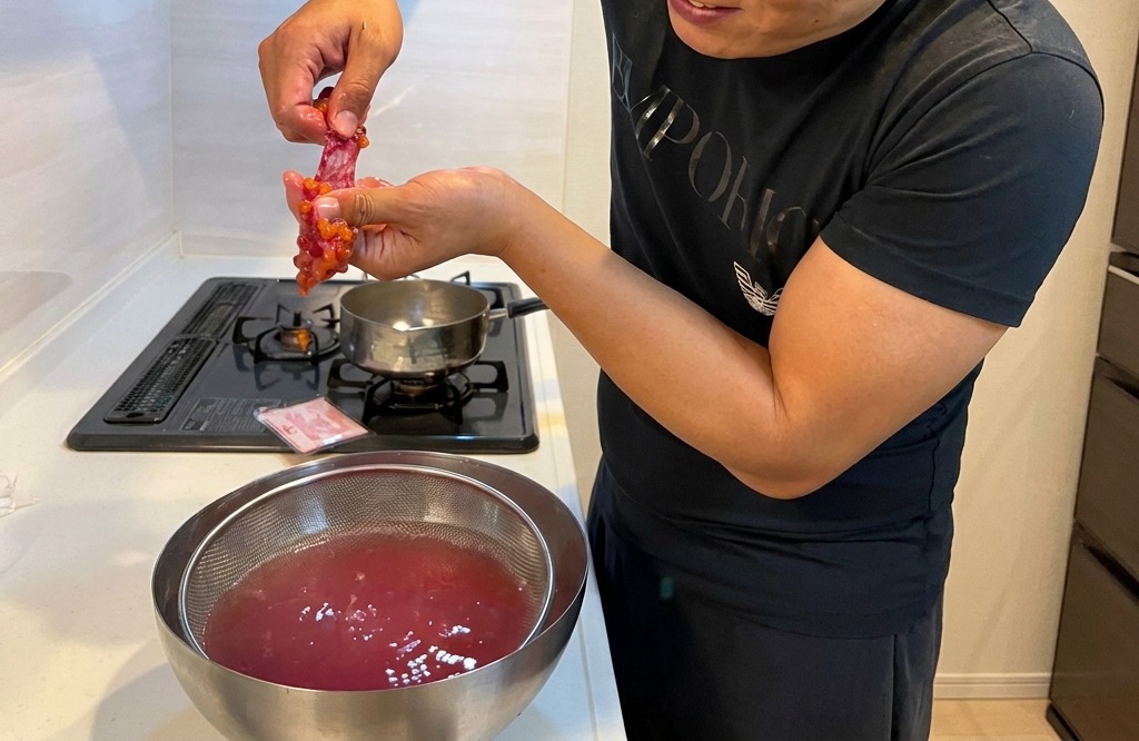 A persons hands gently rubbing a salmon roe sac against a metal sieve submerged in a bowl of warm water to release the individual eggs.
