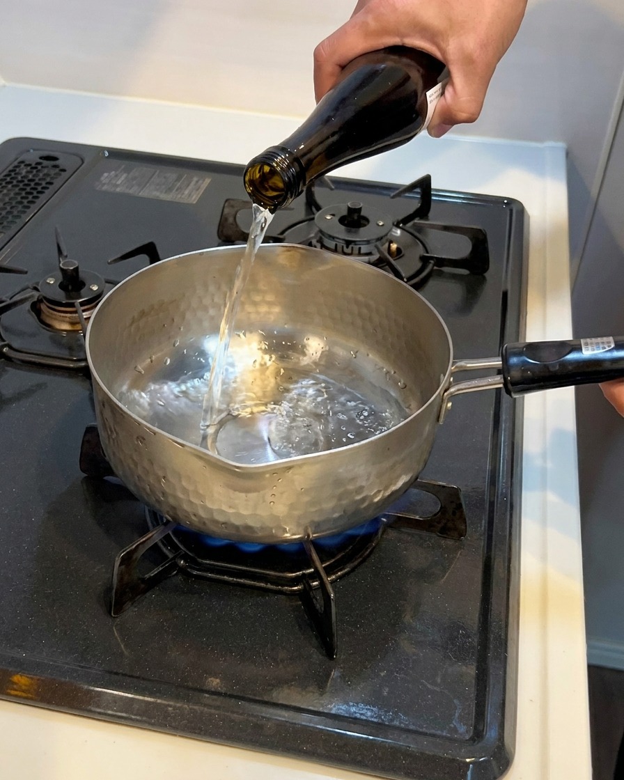 Clear liquid being poured from a dark glass bottle into a small hammered metal pot resting on a gas stovetop.