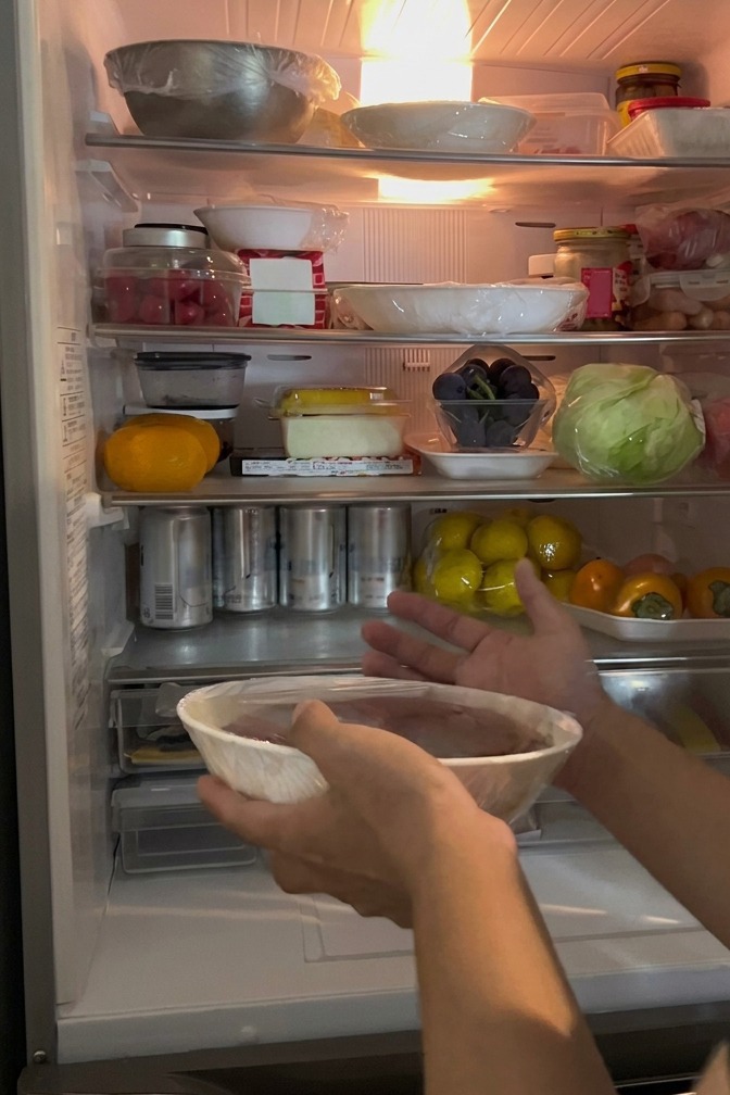 A hand placing a bowl of salmon roe, sealed with clear plastic wrap, onto the middle shelf of a refrigerator filled with various food items.