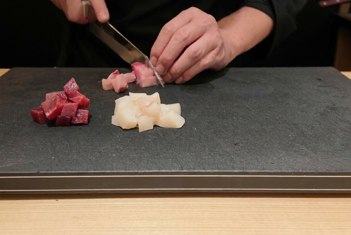 A chef slicing fresh raw tuna and yellowtail into small cubes on a black cutting board.