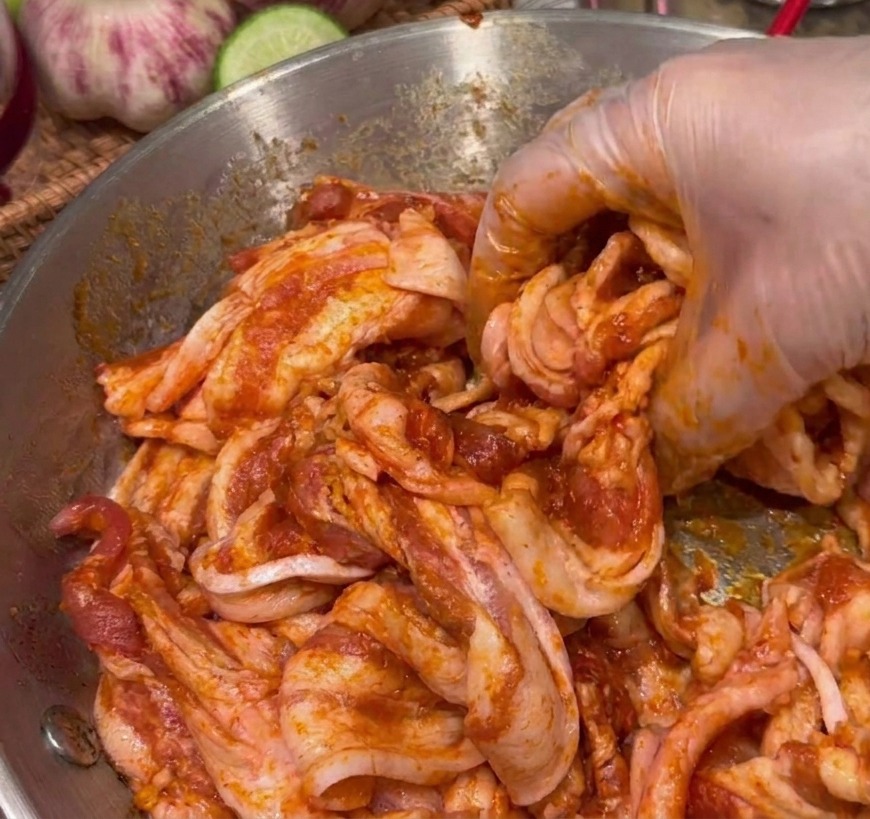 A gloved hand mixing raw pork belly slices with a thick, dark red marinade in a stainless steel bowl.