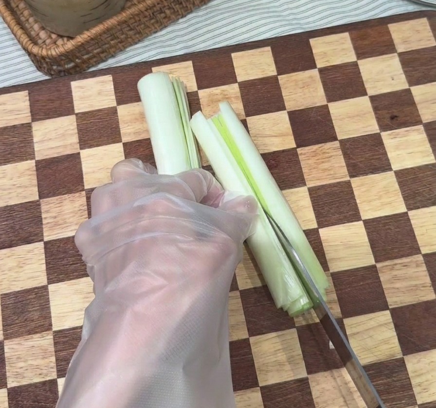 A hand using a chefs knife to slice a white scallion stalk into thin vertical strips on a checkered wooden board.