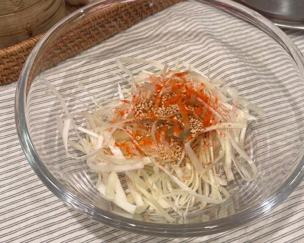 A glass bowl containing white scallion shreds topped with a pile of red chili powder and white sesame seeds.