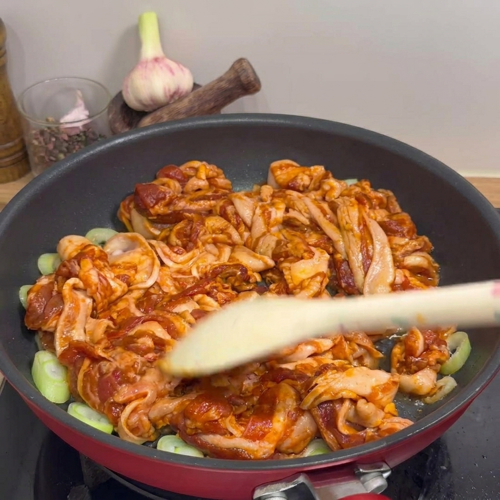 A close-up shot of marinated pork belly strips being stir-fried in a red pan with rings of green onion, using a wooden spatula to turn the meat.