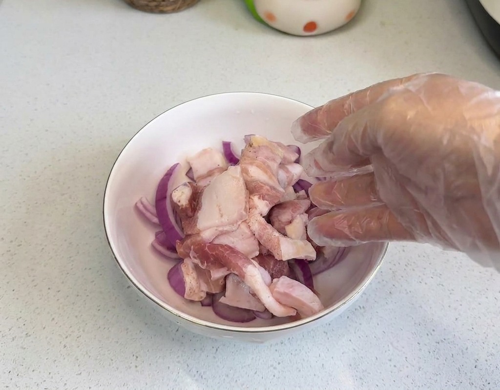 A hand wearing a clear plastic glove placing raw pork belly slices into a white bowl filled with red onion rings.