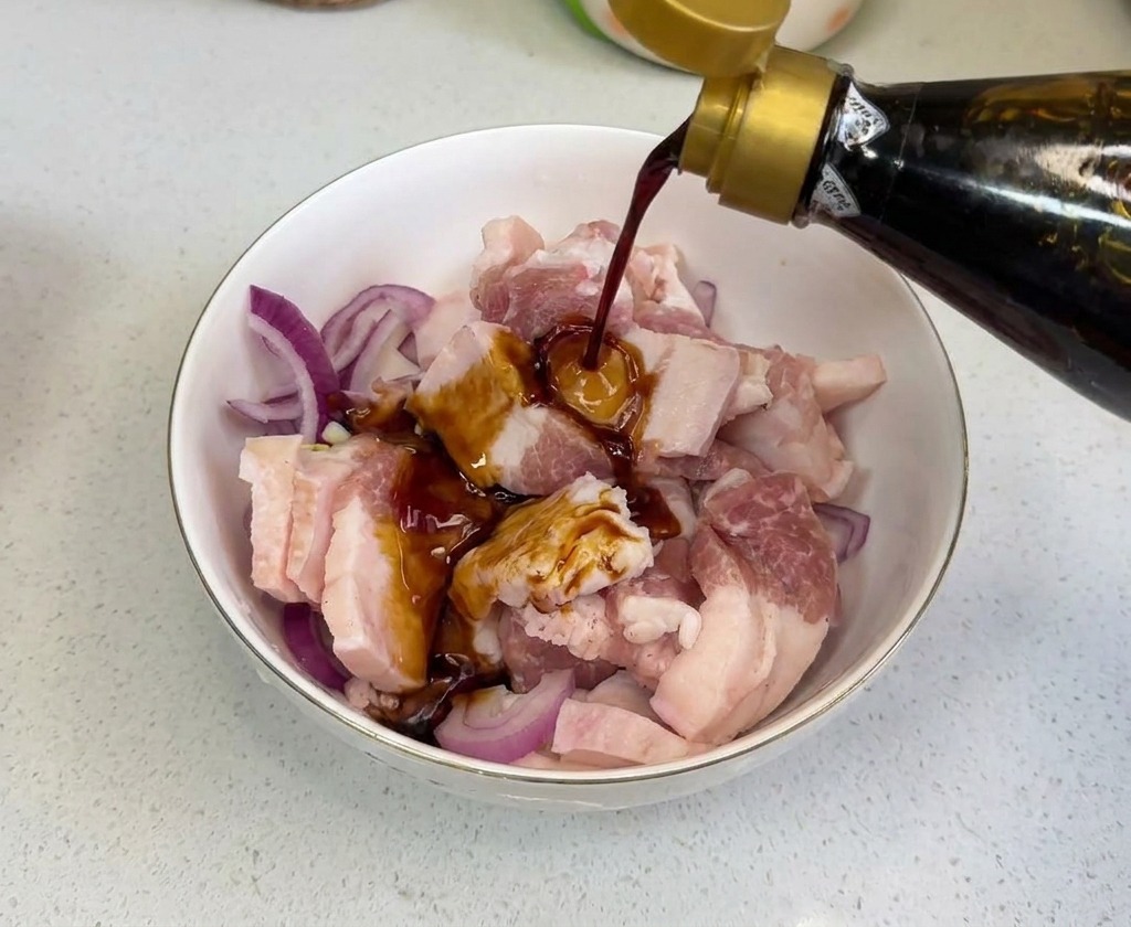 Dark soy sauce being poured from a glass bottle onto raw meat and sliced onions in a mixing bowl.