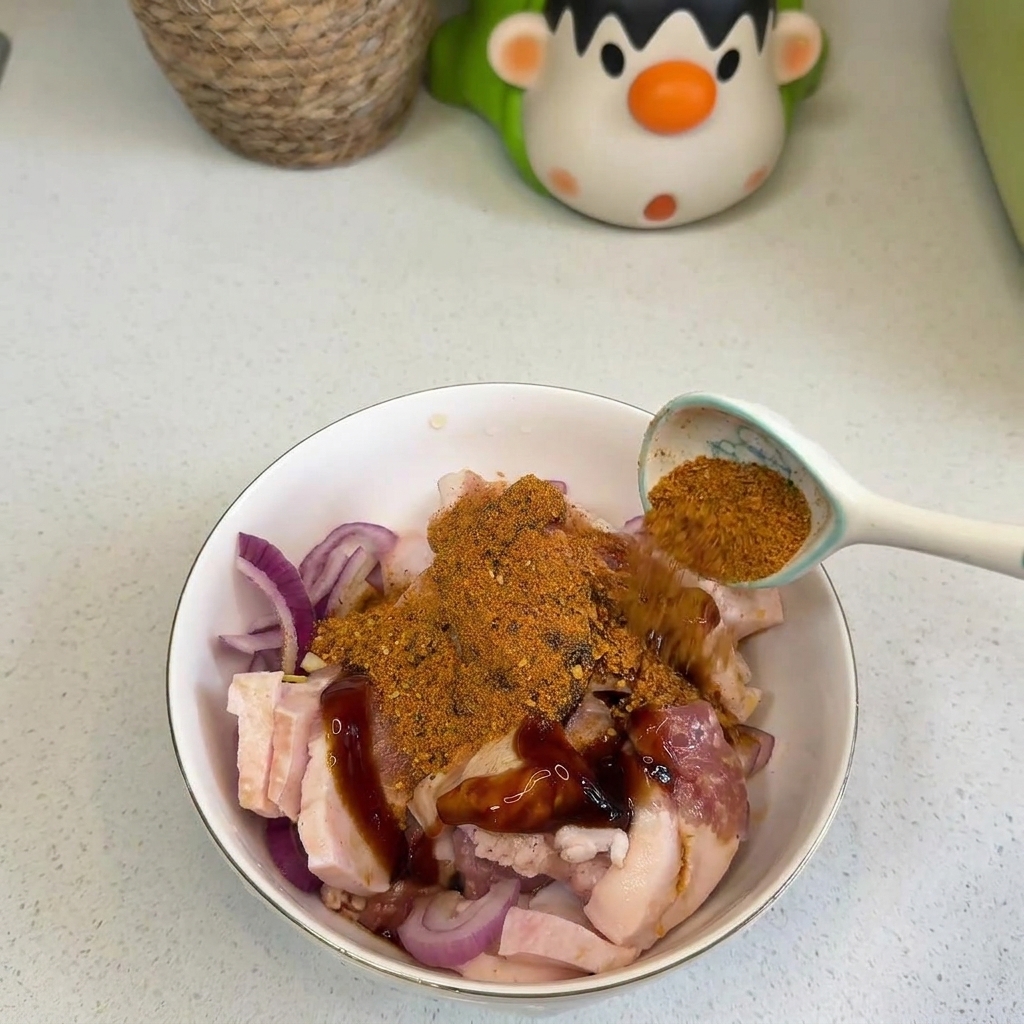 A hand pouring a spoonful of orange BBQ seasoning and red chili powder over raw pork belly slices and red onions in a white bowl.
