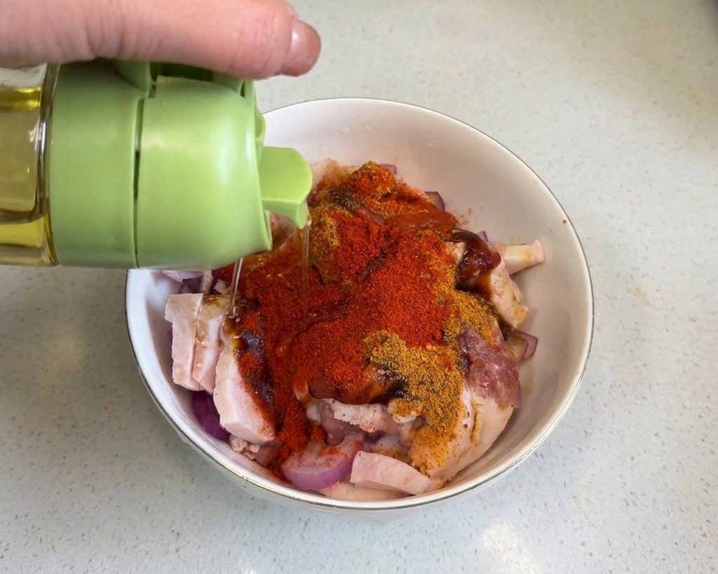Clear cooking oil being drizzled from a green-capped container over pork belly coated in red spice powders.