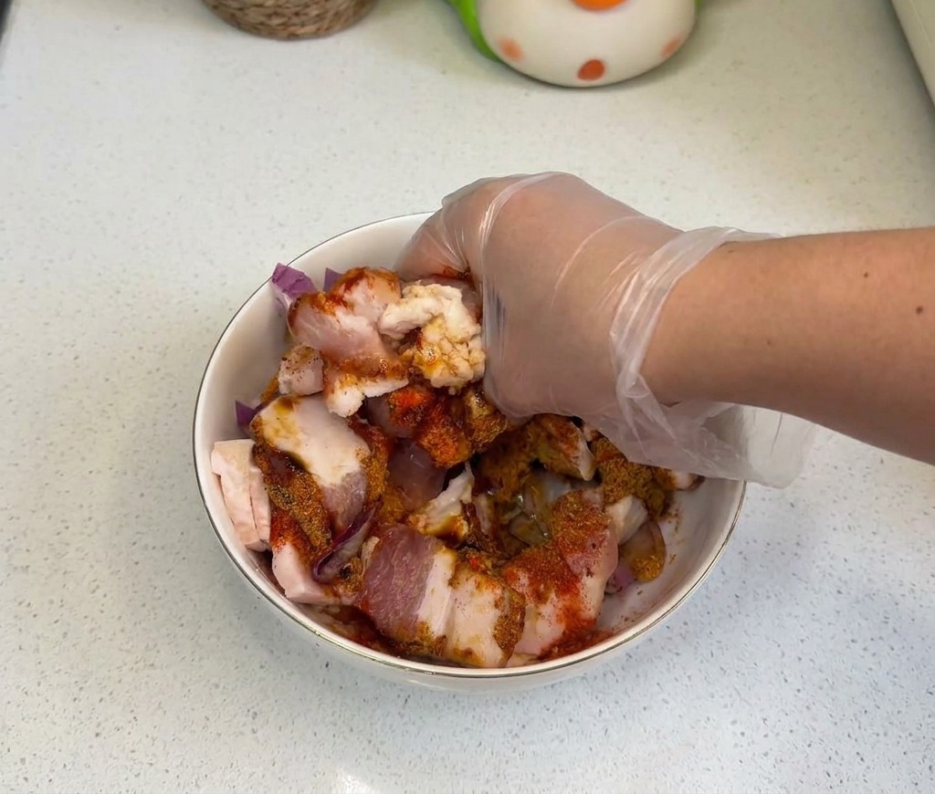 A gloved hand thoroughly mixing together raw pork belly, onions, and seasonings in a white bowl.