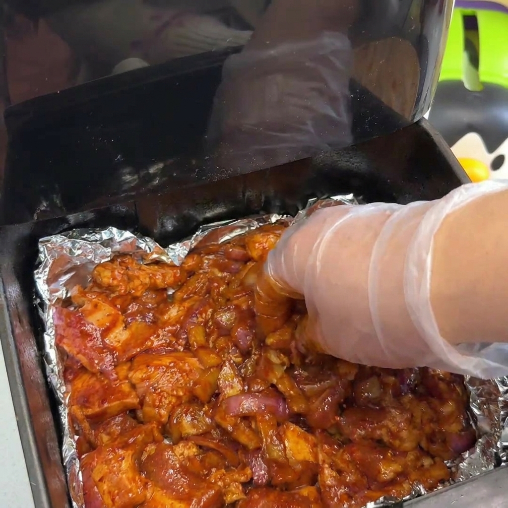 A gloved hand spreading out a thick layer of marinated pork and onions in an air fryer basket.