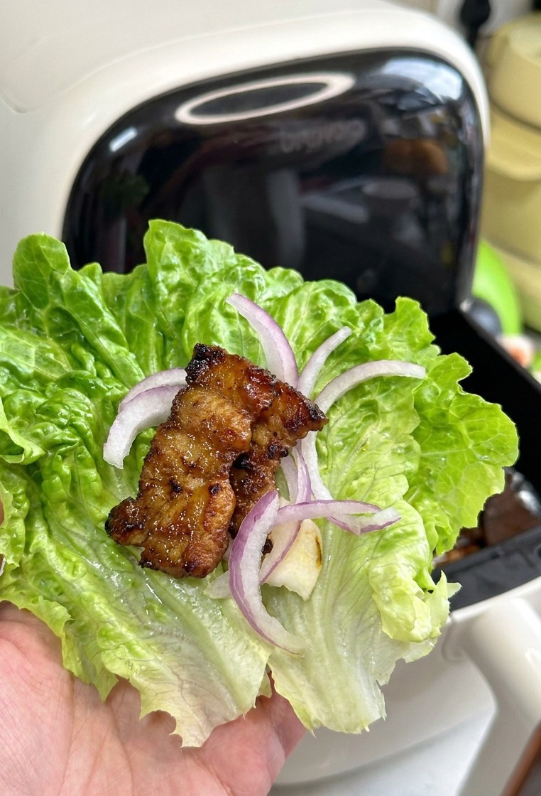 A lettuce leaf held in a hand, containing raw garlic, onion, and a well-browned, glistening piece of roasted pork belly.
